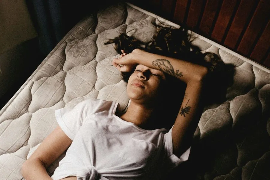 young person on mattress with arm over their forehead looking exhausted