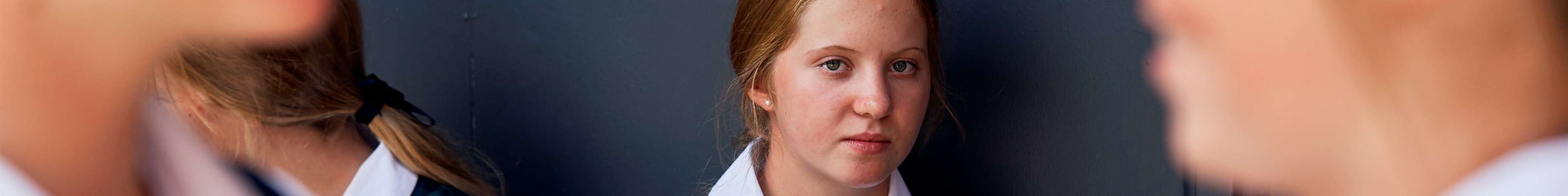 Image of a young teen girl sitting against a wall at school. She's looking at two other teen girls in the foreground, who are talking and laughing.