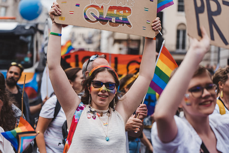 Image of a young woman at a pride parade. She is holding a sign above her head. The word QUEER is visible on the sign, and has been coloured in with colours of the pride flag rainbow.