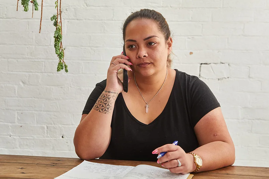 Image of an adult woman sitting at a desk. She has a journal open in front of her and is holding a pen in one hand. Her other hand is holding a mobile phone to her ear.