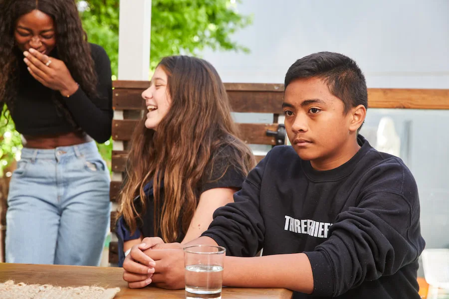 Image of a teen boy sitting at a table, looking sad. Two teen girls are next to him laughing.