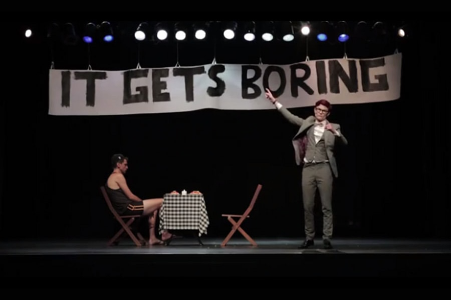 Image of comedian Rhys Nicholson on stage point to a large banner hanging above him that reads IT GETS BORING. There is another man on stage below the banner, sitting at a small table set for two.