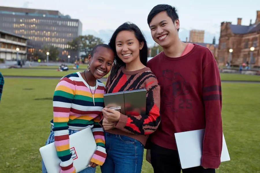 Image of three teenagers standing close together and smiling to the camera. The woman on the left has black skin and shaved hair. She is resting her head on the woman in the middle, who has tanned skin and long black hair. The man on the right has light skin and short black hair.