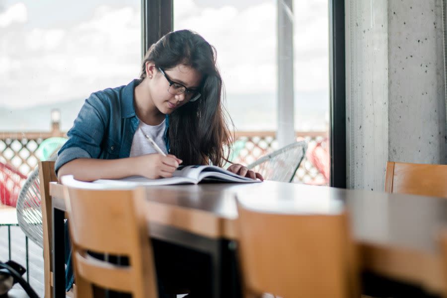 young woman studying at a long table in denim shirt over white tshirt with long black hair and black rimmed glasses.