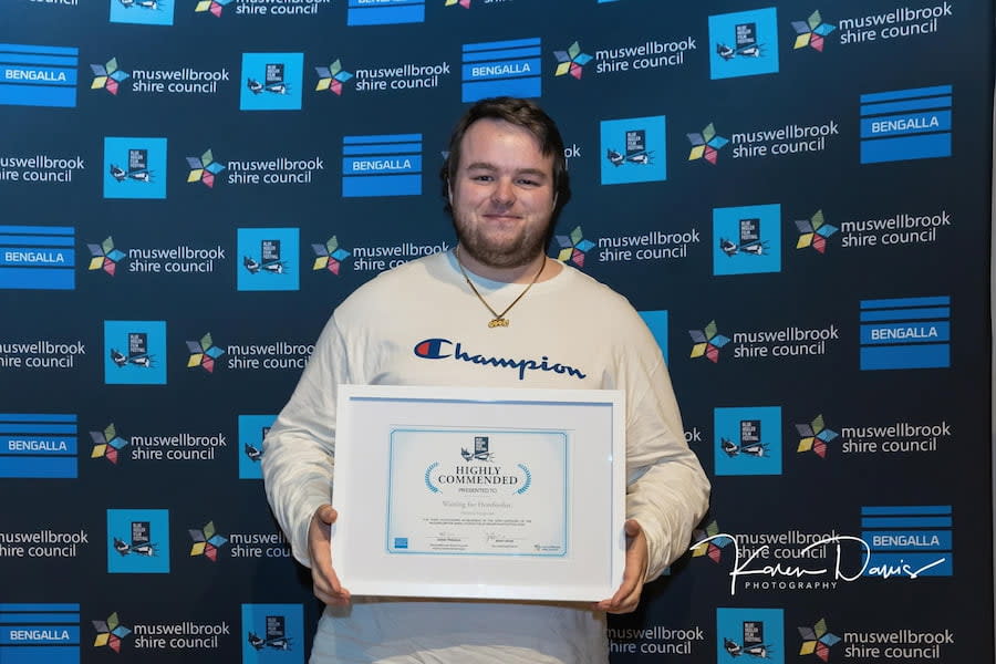 Dominic, the article author, stands in front of a photo wall that is branded with Muswellbrook Shire Council and film festival logos. He holds an award that says 'highly commended' and is smiling.