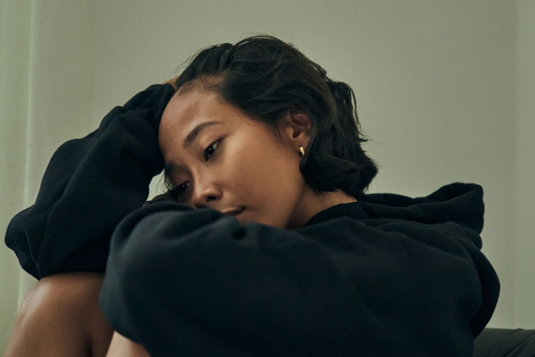 young girl in a black hoodie sitting with one arm folded over bent up knees and head resting in other hand looking sad.