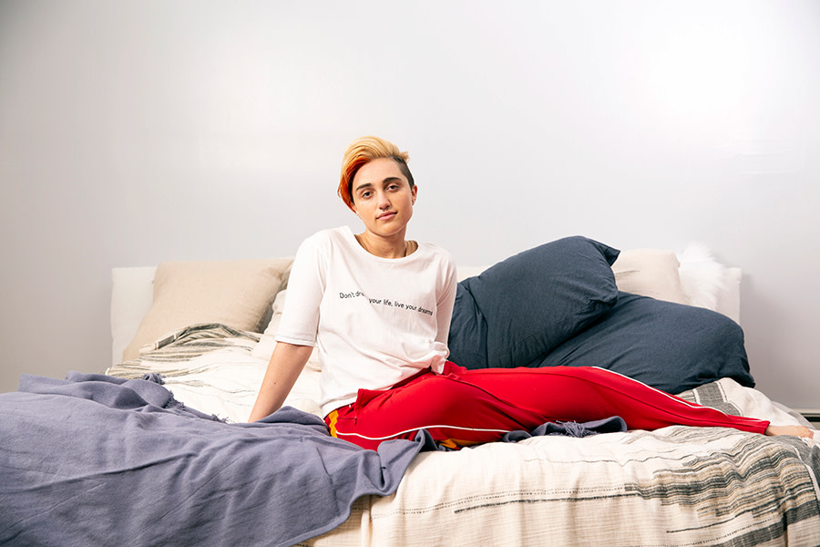 Image of a transmasc gender-nonconforming person sitting on a bed. They are looking at the camera with a very small smile.