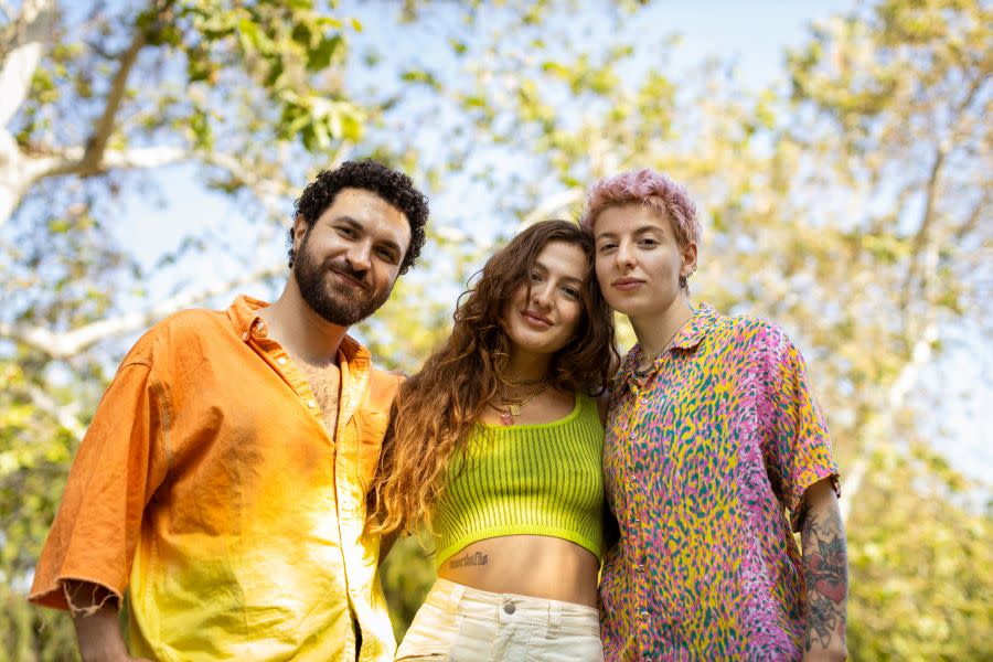 Three friends standing shoulder to shoulder in colourful clothing on a sunny day looking supportive.