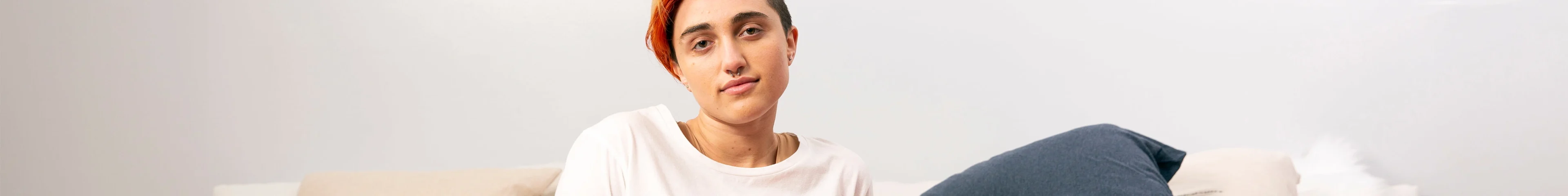 Image of a young gender non-conforming person sitting on their bed and softly smiling to the camera.