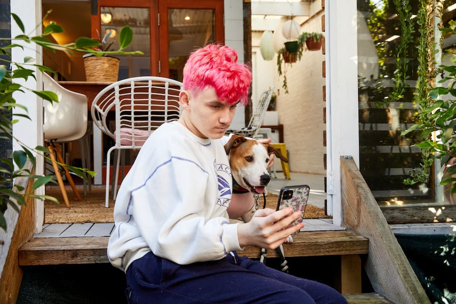 Image of a pink-haired teen sitting on the steps outside their home and looking at their phone. Their service dog sits at their side.