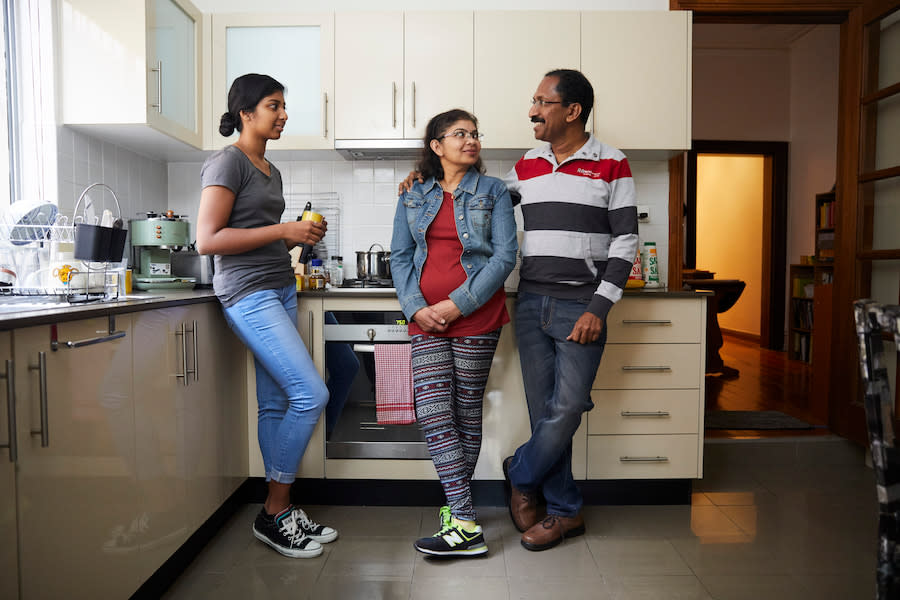 Image of a teen leaning on a kitchen counter looking at her parents, who are standing close together and smiling.