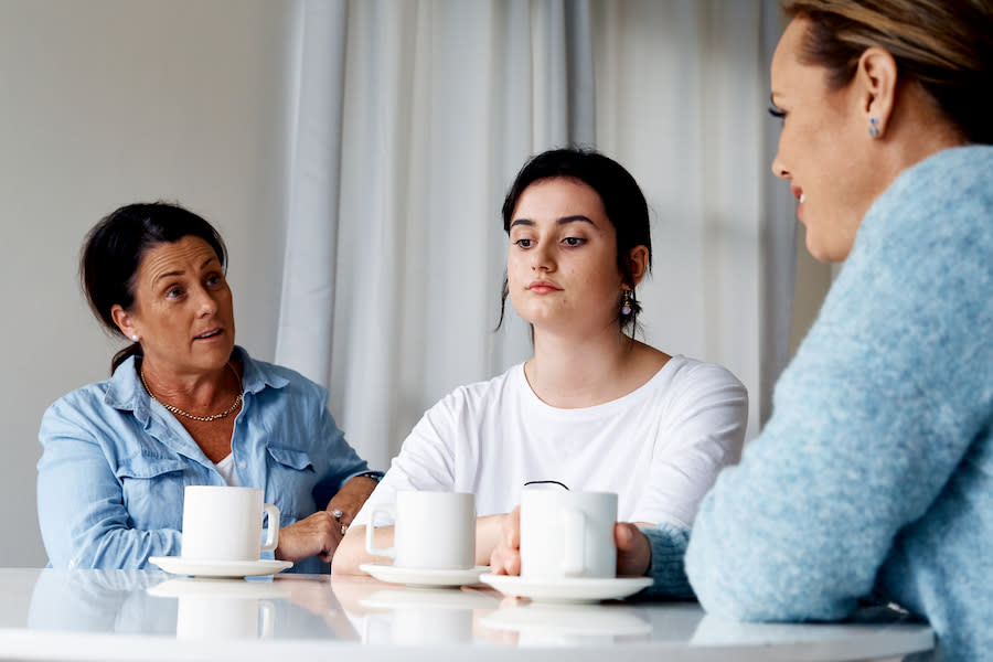 Image of a teen girl sitting between her two mothers and looking down at the table in front of her. Her parents are talking to each other over her.