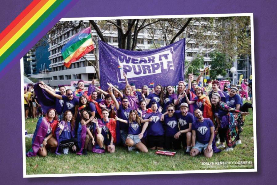 Image of a large group of people in a parking posing for a photo underneath a banner that reads WEAR IT PURPLE. They are all dressed in purple t-shirts. A rainbow pride flag is flying above them.