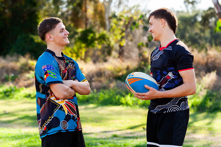 Image of two teen boys chatting outside. They are both wearing sports jerseys and one is holding a football.
