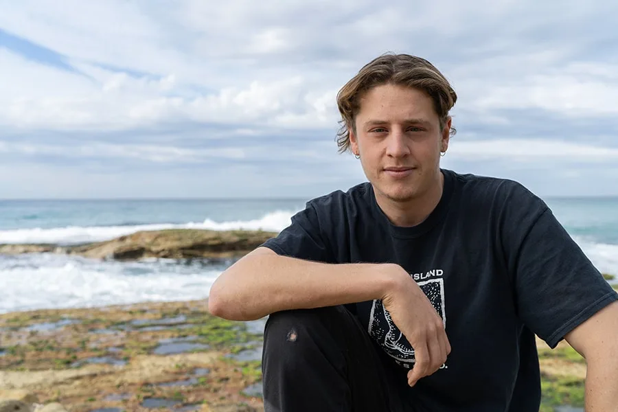 Christo guy sitting casually with beach foreshore behind him