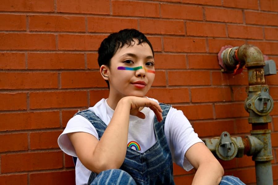 young non-binary person with short brown hair and a rainbow strip painted their face sitting against an orange brick wall.
