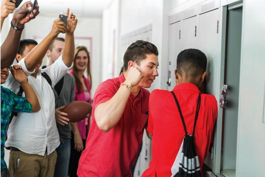 Image of a teen boy physically intimidating another teen. Several students are watching this interaction, and filming it on their phones.