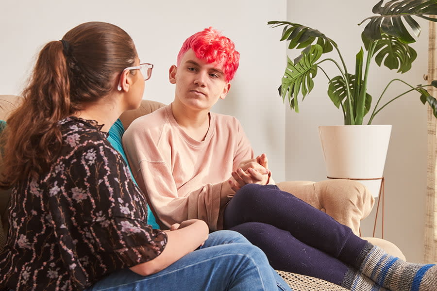 Image of a teen boy with bright pink hair sitting on a lounge, speaking with a friend.