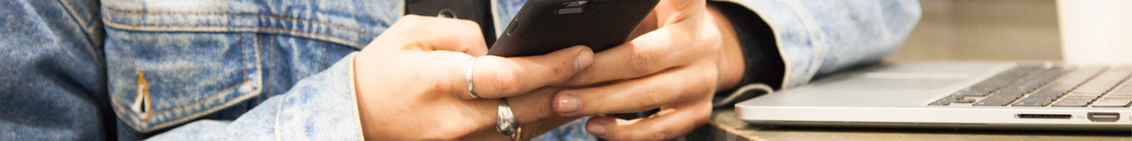 Image of a teen holing a phone, sitting on a desk next to a laptop.