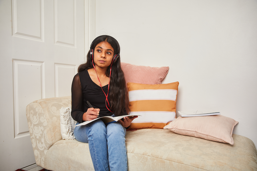 Image of young woman on a lounge with headphones on, writing in a journal.