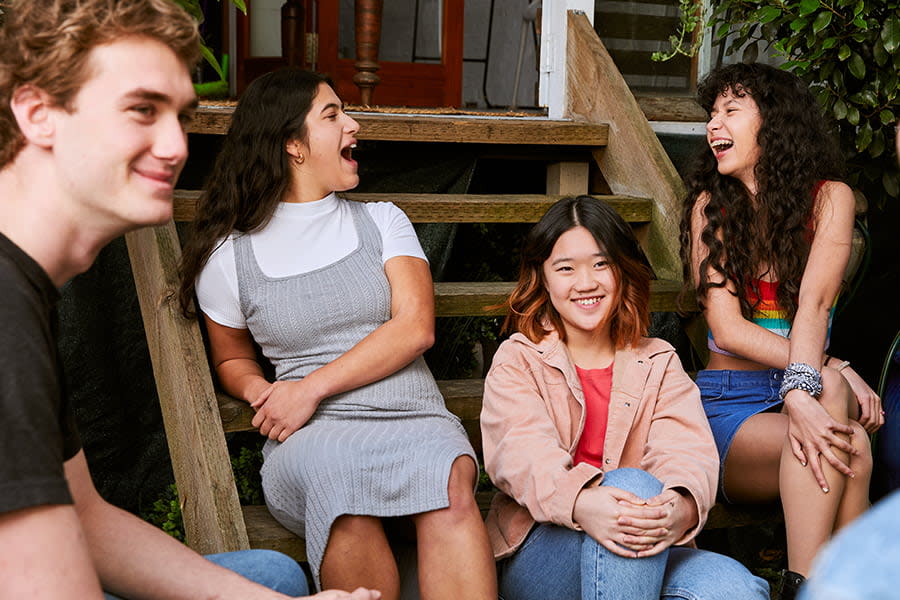 Image of four teens sitting together outside. Two are looking at a person off camera and smiling, and two are looking at each other and laughing.