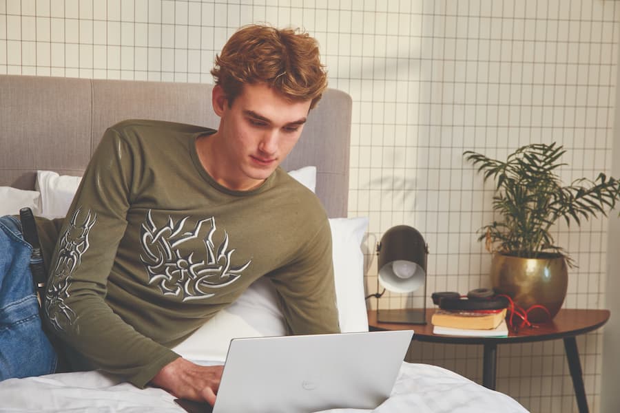 Image of a late teenage boy lying on a bed and writing on a laptop.