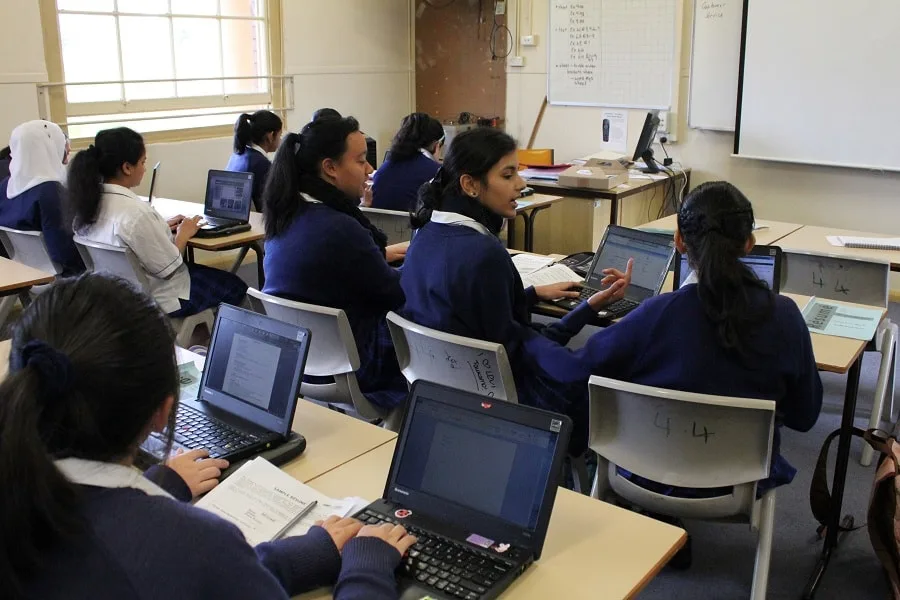 Image of high schoolers in a classroom chatting. They each have a laptop open in front of them on their desks.