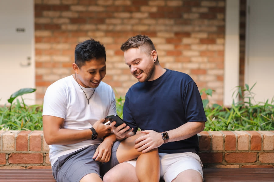 Image of two young men sitting outside. They are both smiling as they look down at one of their phones. Their legs are intertwined, and one man has his hand rested on the others knee.