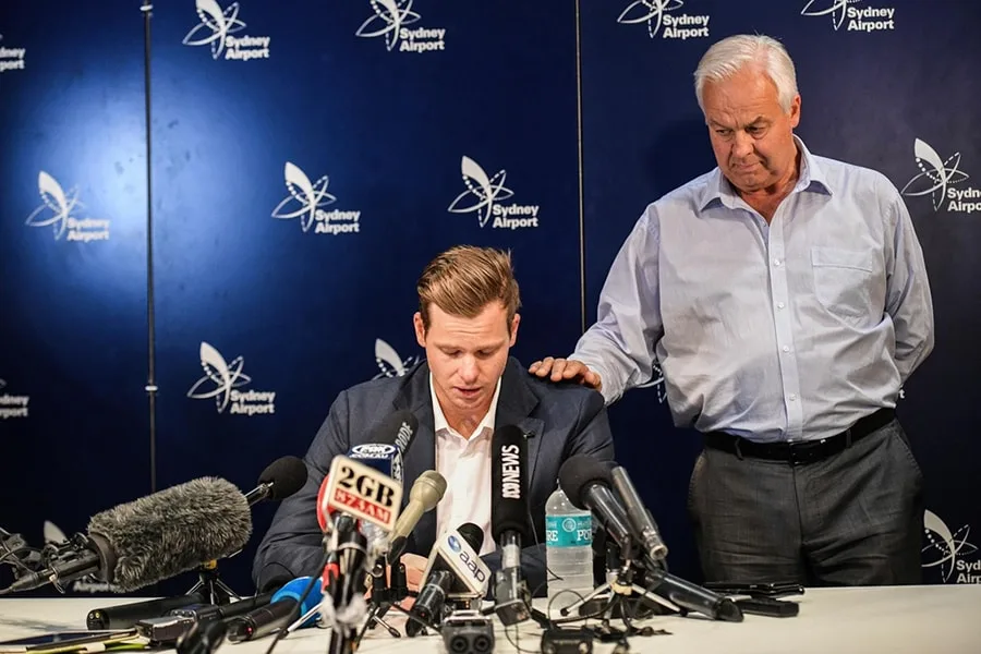 steve smith cricketer looking down speaking into microphones with man next to him with his hand on s