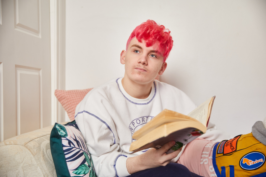 A teenager with pink hair is sitting on the couch with a book. The book is open but they are looking at something else in the distance.
