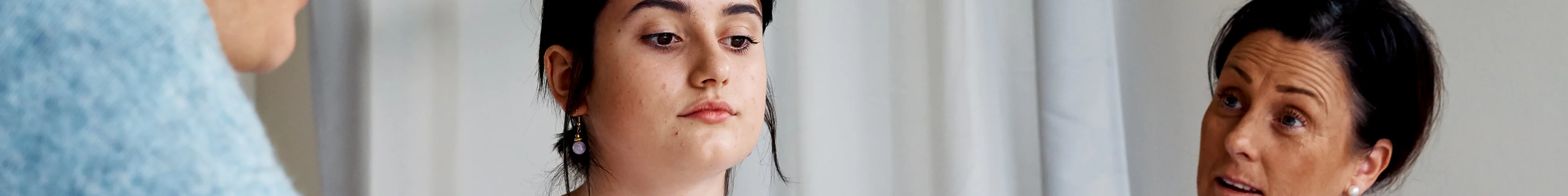 Image of a teen girl sitting between her two mothers and looking down at the table in front of her. Her parents are talking to each other over her.