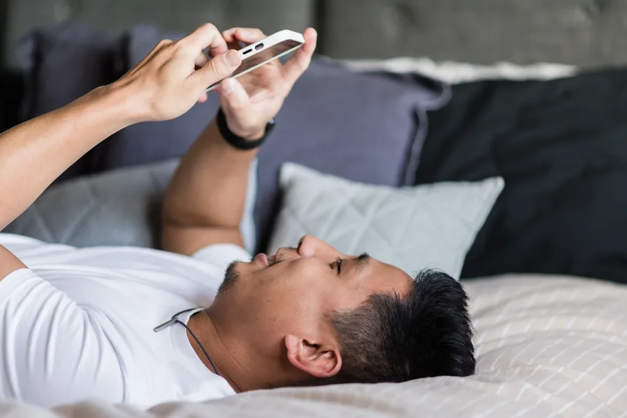 A young man lying on his bed holding his phone above his face and smiling.