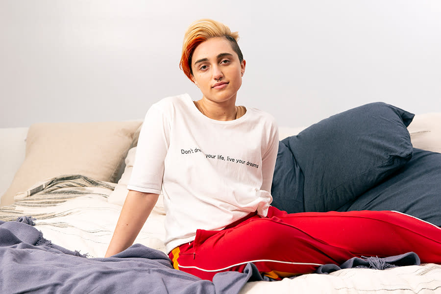 Image of a young gender non-conforming person sitting on their bed and softly smiling to the camera.