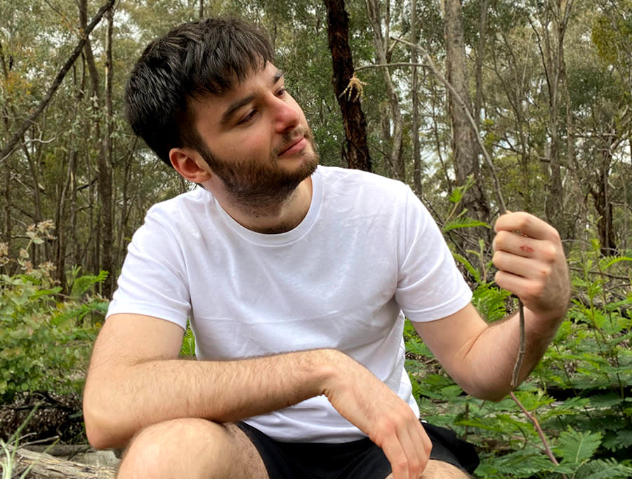Image of Aidan, a young man with light skin, short black hair and a beard. He is sitting outside in nature, staring peacefully at a plant.