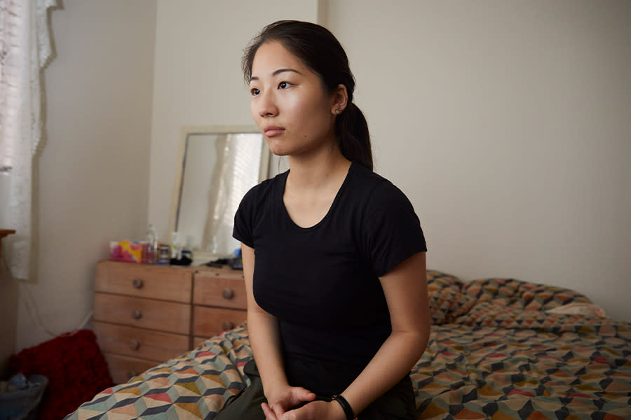 Image of a teen girl sitting on her bed. She is looking away from the camera, and appears anxious. Her hands are intertwined and resting in her lap.