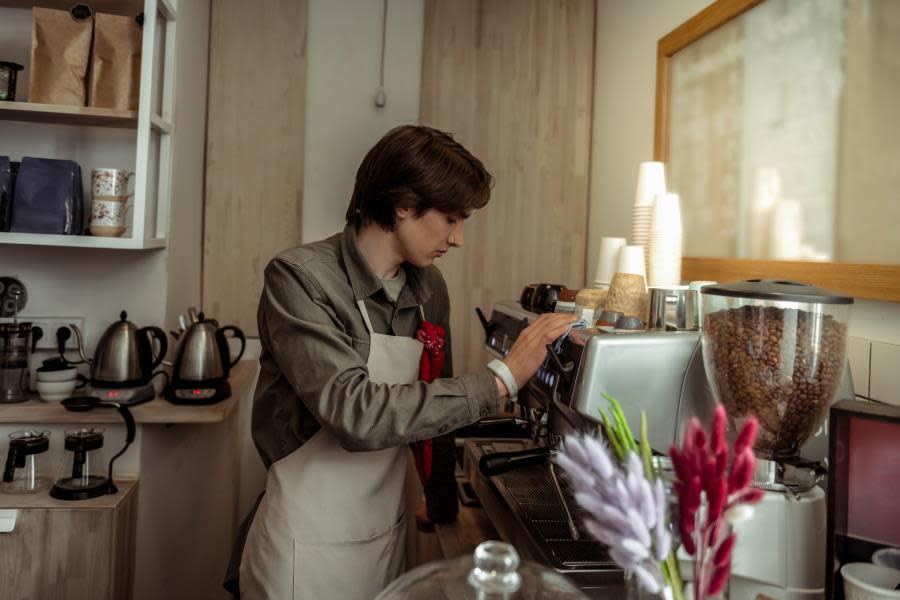 young male barista with a nose piercing wearing an apron wipes down a commercial espresso machine