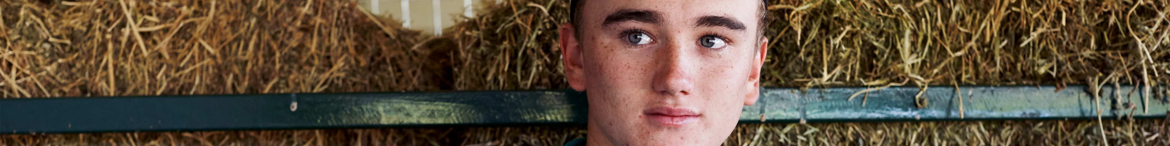 Image of a teen boy standing in front of some hay bales. He is looking away from the camera and isn't smiling.