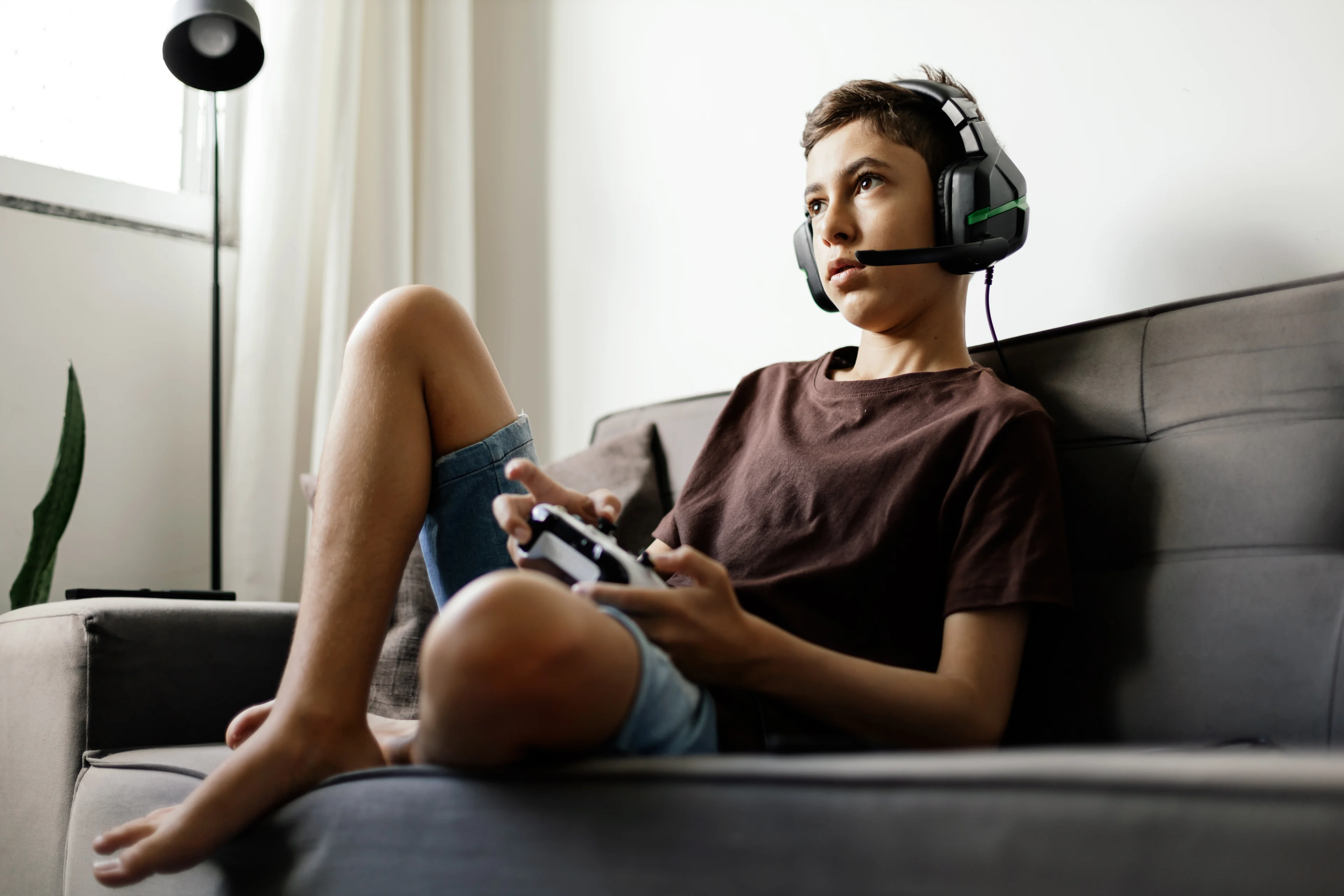 A teenager wearing a headset sits on the couch while playing a video game.