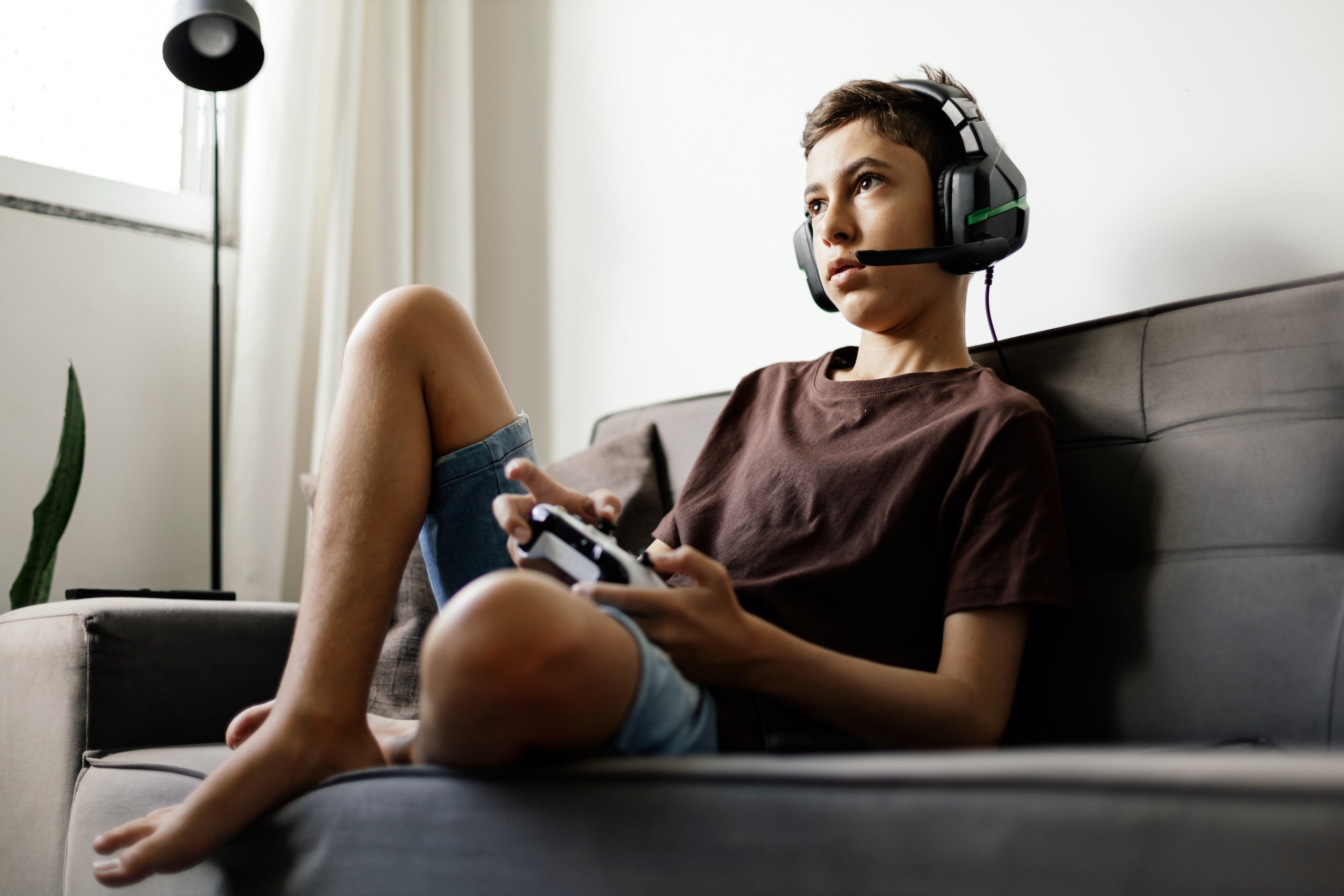 A teenager wearing a headset sits on the couch while playing a video game.