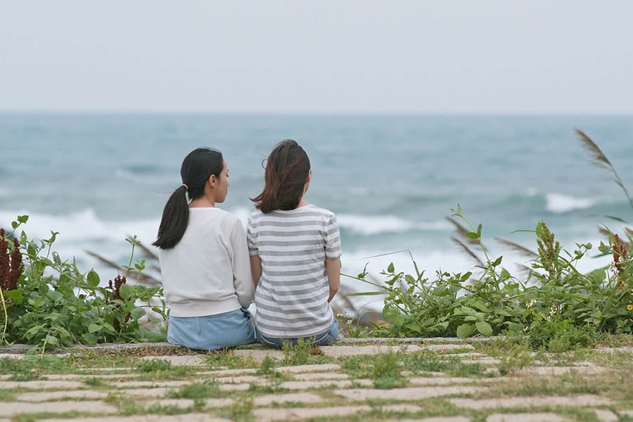 two young women sit facing the ocean