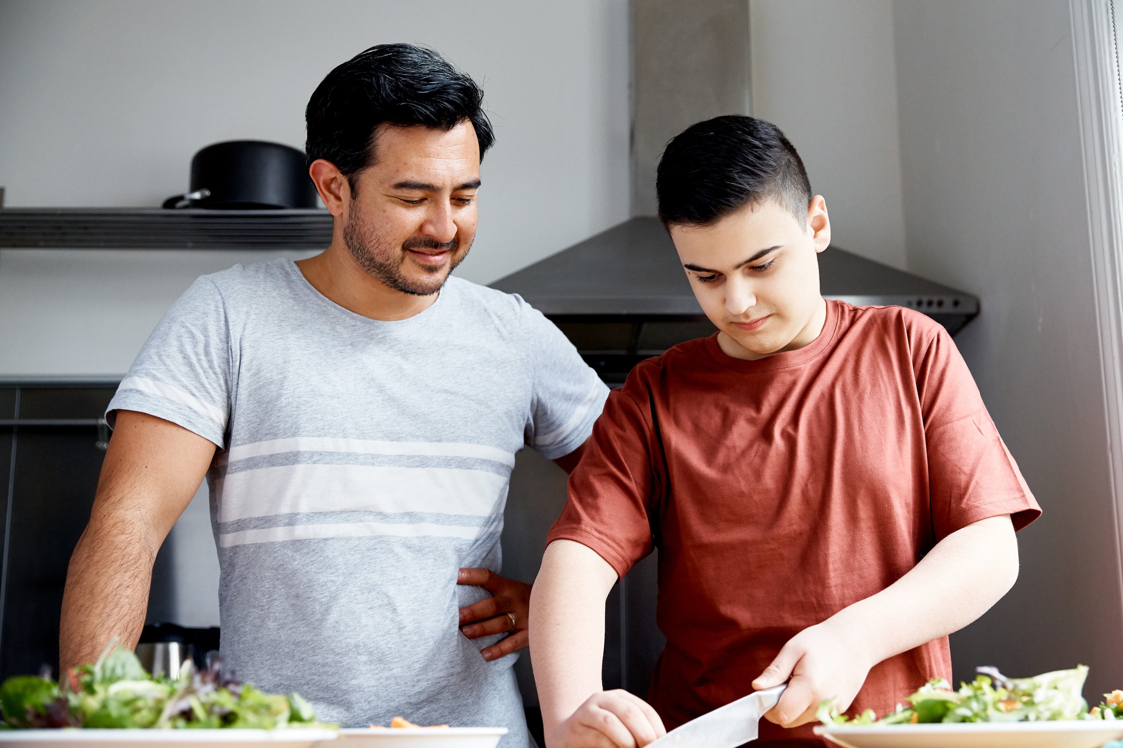 A dad stands next to his son in the kitchen and watches on while the son chops some ingredients. There are two plates with salad in front of them.