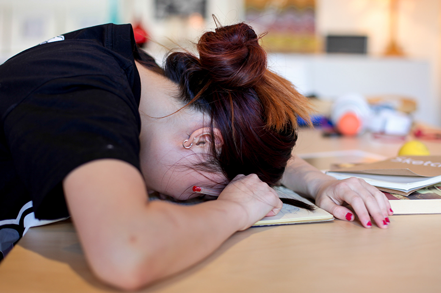 Image of a girl resting her head on a desk. She's surrounded by study notes.
