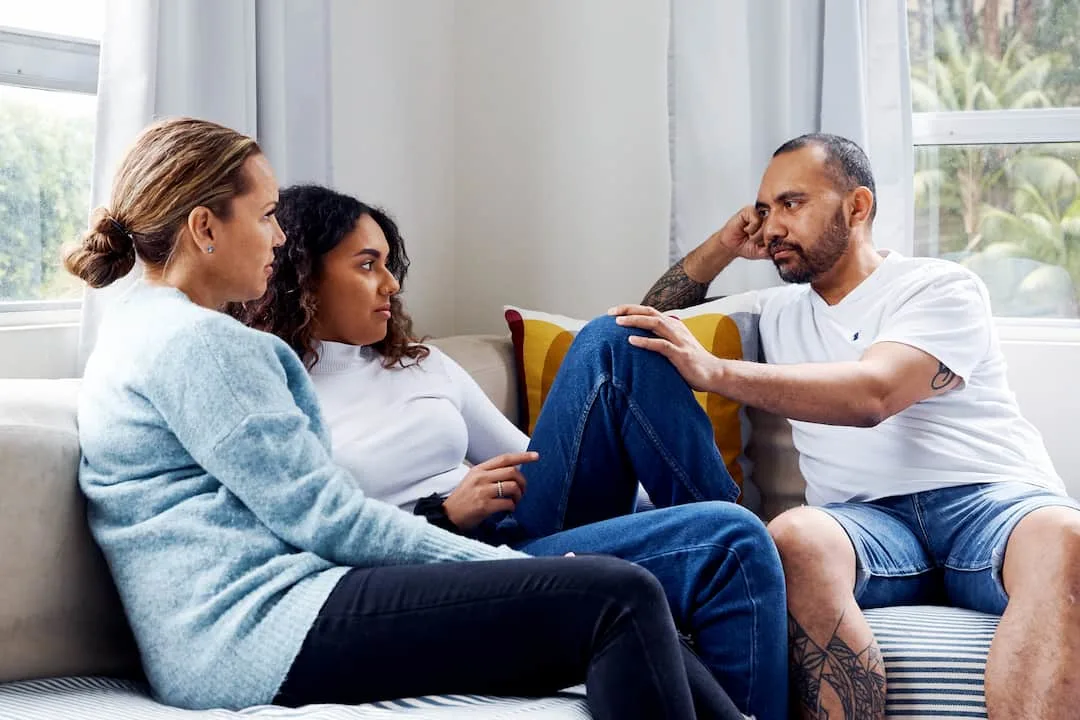 A teen girl sitting between her two parents. Her father is looking at her with concern, and is resting his hand on her knee in comfort.