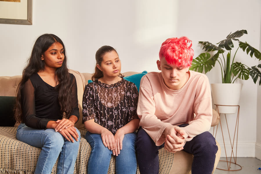 Image of three young people sitting on a lounge. The two girls on the left have neutral expressions and are looking at the man on the end, who is sitting with his head hanging down and his hands crossed on in his lap. He looks upset.