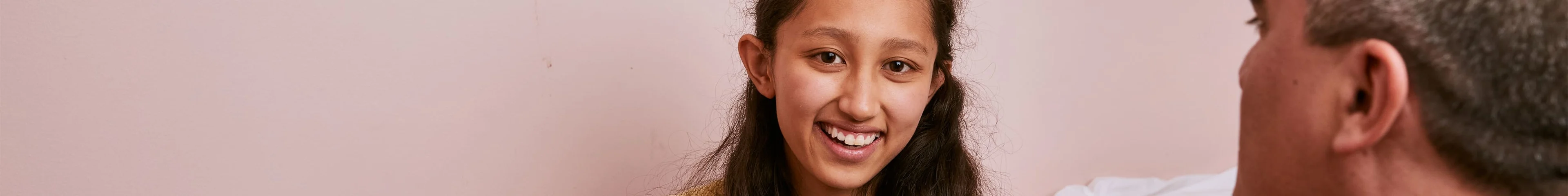 Image of a young teen sitting on her bed, smiling to camera. Her Dad sits across from her, and is speaking to her.