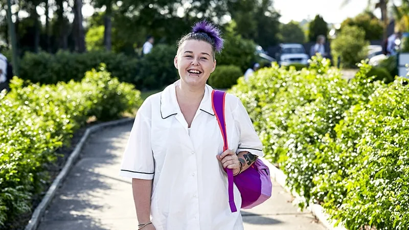 a girl in school uniform walking down a path smiling with a backpack over her shoulder