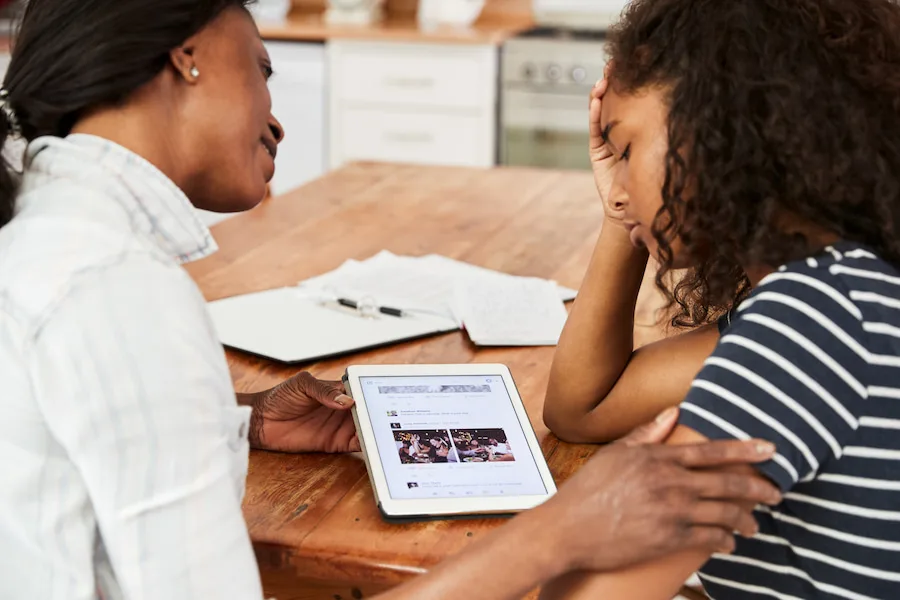 Image of a mother and daughter sitting at a dining table looking at an iPad. The teen daughter looks stressed, and the mother is comforting her.