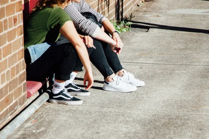 Two girls sitting next to each other on the sidewalk talking.