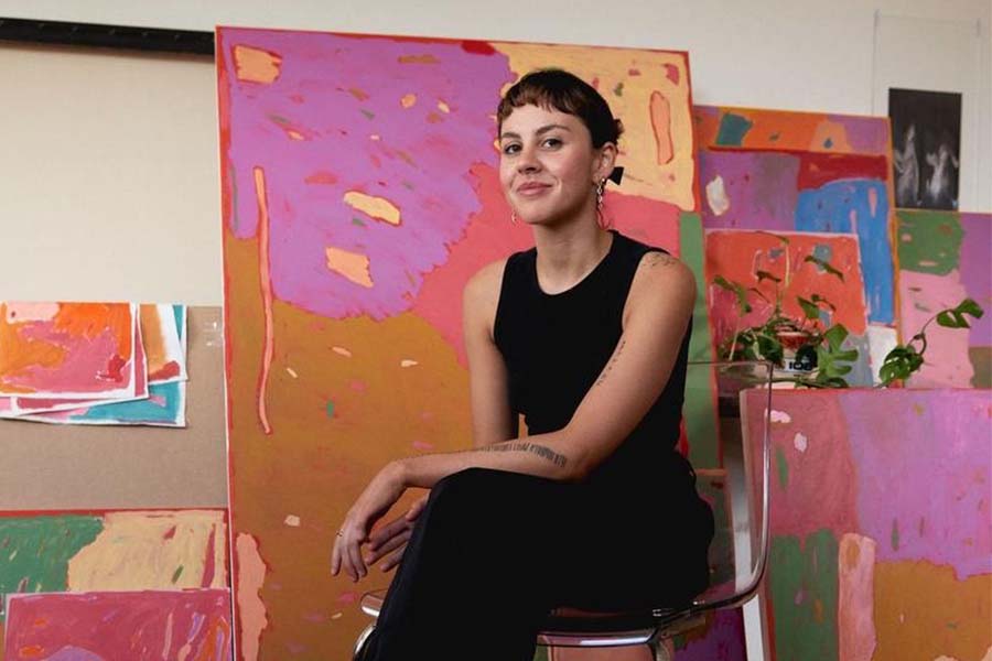 a woman wearing all black clothing sits on a chair in front of a collection of her paintings1