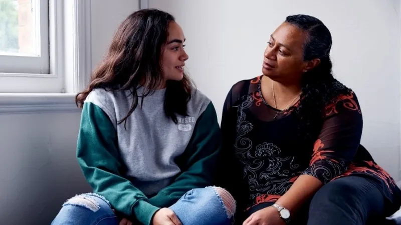 A young teen girl with long dark hair (left) looks happy as she sits on her bed talking to her mother (right).